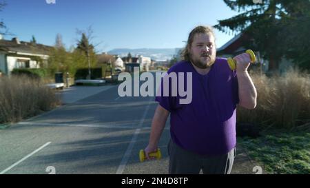 A young fat man getting back into shape building strength. Motivational concept of an overweight person listing weight dumbbells standing outside Stock Photo