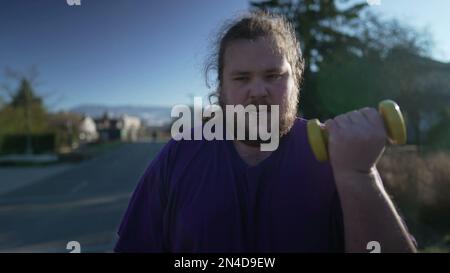 One disciplined young fat man lifting dumbbells outdoors. Motivational concept of an overweight person getting back into shape Stock Photo