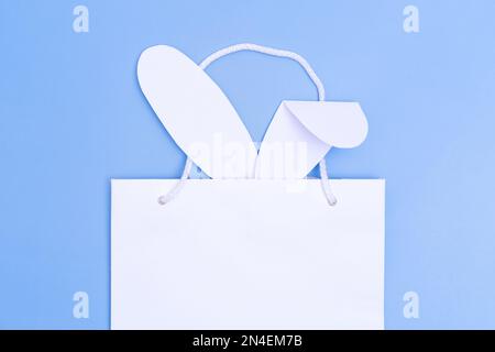 Shopping for Easter. White paper shopping bag with white bunny ears on blue background. Concept Easter gifts, preparation, holiday surprise. Stock Photo