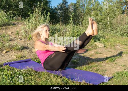 An elderly woman lifting her legs up, practicing yoga on the lake shore in summer. Meditation. Sports in old age. Yoga on the shore. Summer sports Stock Photo