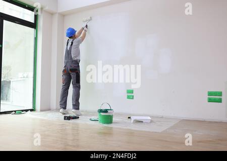 Decorator in hard hat paints apartment wall with roller brush. Stock Photo