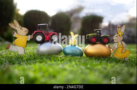 two models of toy tractors stand on painted multi-colored eggs that lie on the grass. concept of commercial easter greetings for farms, agribusiness. Stock Photo