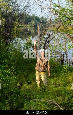 Yakut Asian girl tourist dressed in protective color clothes waving his hand standing on the shore of overgrown wild lake in the forest. Stock Photo