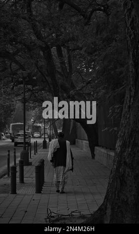 A vertical shot of a man in traditional clothing walking on a street in grayscale Stock Photo