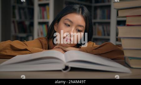 Bored sad tired lazy young korean woman read boring book lying on desk in university library unmotivated female student doing homework preparing for Stock Photo