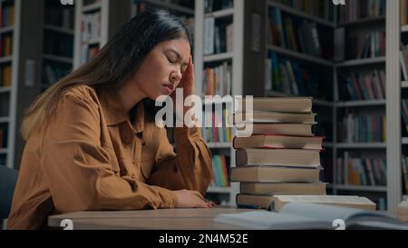 Tired unwell young korean woman student preparing for exam in university library feeling headache suffering from study overload having painful feel Stock Photo