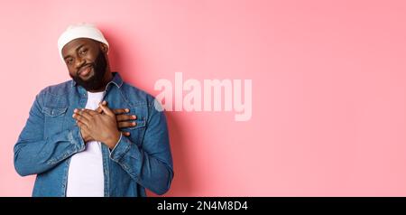 Thankful and touched Black guy saying thank you, smiling adorable and holding hands on heart, appreciate nice gesture, standing over pink background Stock Photo