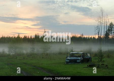 The rusty remains of the Soviet iron car lie in the forest near the road in a fog strip at sunset in the North of the Yakut taiga. Stock Photo