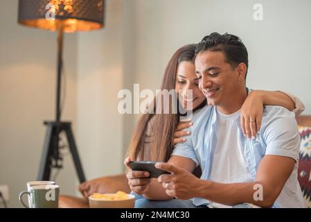 Smiling millennial couple sit on sofa in living room have fun using modern smartphone devices together, happy young husband and wife laugh relax at ho Stock Photo