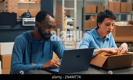 Diverse partners checking quality of goods before packing them in cardboard boxes, putting merchandise to ship. Young entrepreneurs working together on shipment, retail distribution. Stock Photo
