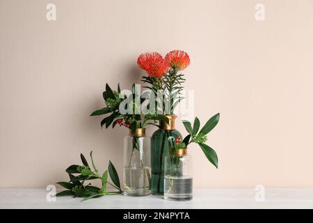 Vases with beautiful flowers and branches on white wooden table near beige wall Stock Photo