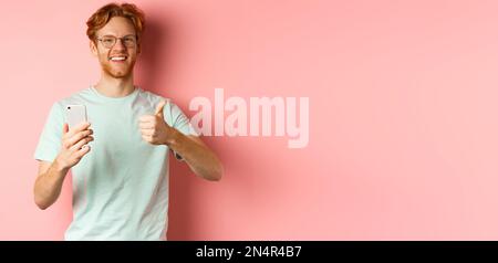 Happy young man with red messy hair, showing thumbs-up while using mobile phone, smiling and praising application, standing over pink background Stock Photo