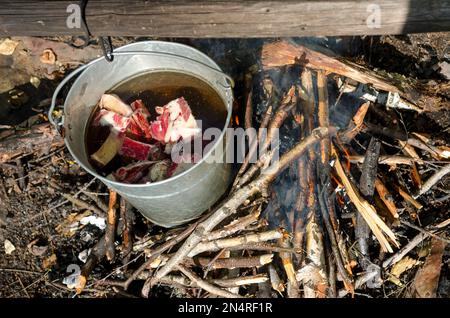 Pieces of meat swimming in a bucket on a camp fire in the wild nature of the North next to the fresh wood. Food at rest. Stock Photo