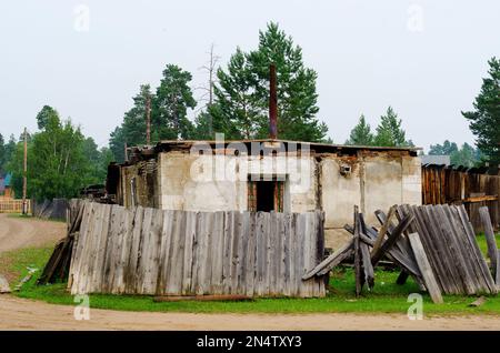An old, rickety wooden barn stands with an open door behind a fallen fence in the Northern village of Yakutia against a forest of pines. Stock Photo