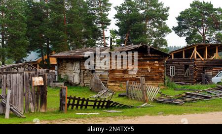 An old, abandoned wooden barn stands behind a fallen fence in the Northern village of Yakutia against a forest of pines. Stock Photo