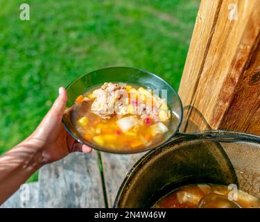The hands of the girl cook outdoors puts soup with meat and vegetables and potatoes, beet borscht from a large cauldron pan on the wooden floor, in th Stock Photo