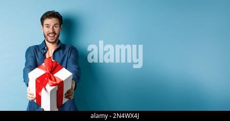 Valentines day. Excited handsome man giving gift box and wishing happy holiday. Romantic guy extending hands with present, standing againt blue Stock Photo