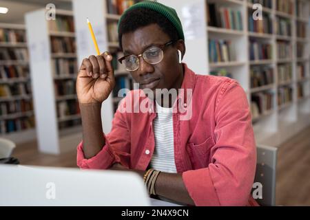 Focused African American student guy in glasses studying sitting in library watching online lesson Stock Photo