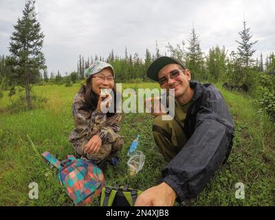 Russian man with glasses making a selfie with a happy Asian girl Yakut for a picnic with sandwiches in the Northern taiga wild forest. Stock Photo