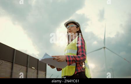 Asian woman engineer working and holding the report at wind turbine farm power generator station. Stock Photo