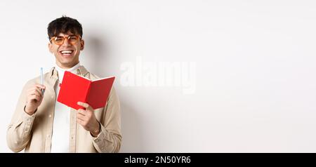 Cheerful young man in glasses laughing and taking notes, writing down in planner, holding pen and diary, standing on white background Stock Photo