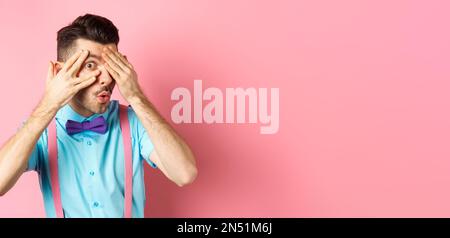 Intrigued funny guy in bow-tie, peeking through fingers at something interesting, checking out something cool, standing on pink background Stock Photo