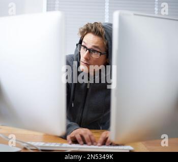 Works squeezing every bit of energy. A young hacker wearing a hoodie working on dual screens. Stock Photo
