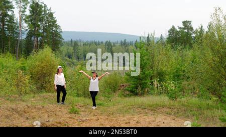 Two joyful girls friends of an Asian Yakut stand on a mountain slope in the taiga forest of Yakutia. Stock Photo