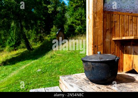 A large, black from soot, the cauldron, the pot stands on the edge of a wooden house a bright day amid toilet in the woods and grass on the field. Stock Photo