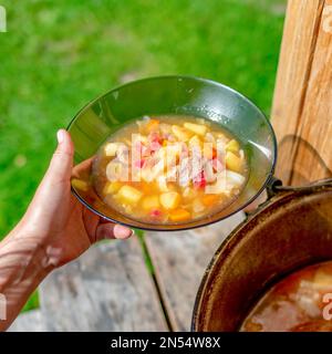 The girl's hands outdoors in the field holding a transparent plate of soup borscht with meat and vegetables on the background of a cauldron with food Stock Photo
