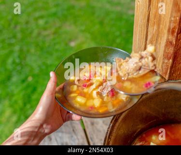 Hands girl cooks outdoors puts soup with meat and vegetables and potatoes, beet borscht from a large cauldron pan on the wooden floor, in a house next Stock Photo
