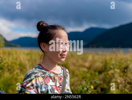 A young Asian girl in glasses looks into the distance against the background of mountains and lake Teletskoye in Altai. Stock Photo
