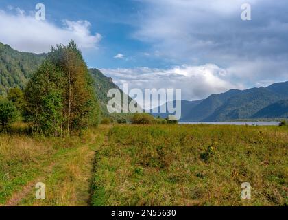 Landscape of the road among the fields near the Teletskoye lake in the Altai mountains with trees and silhouettes of tourists in the distance. Stock Photo