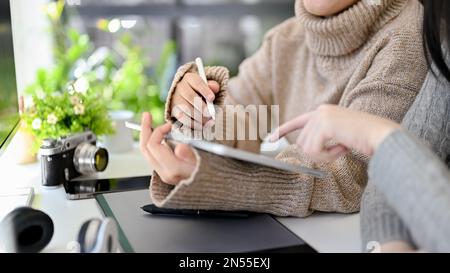 Creative Asian female graphic designer in cozy sweater using digital tablet and discussing work with her colleague in the office. cropped shot Stock Photo