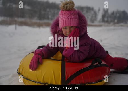 Cute little girl enjoying sledding in the snow close-up. Children sledges. The kid is riding a sleigh in colorful fashionable clothes. Stock Photo