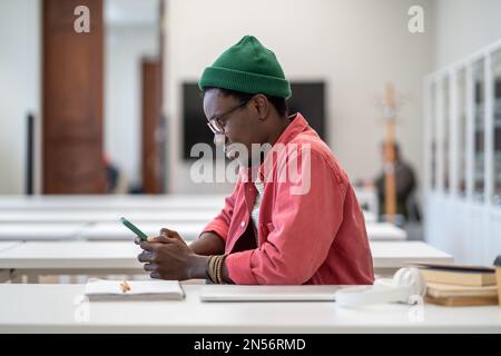 Smiling African guy student using smartphone during study break, sitting at desk in library Stock Photo