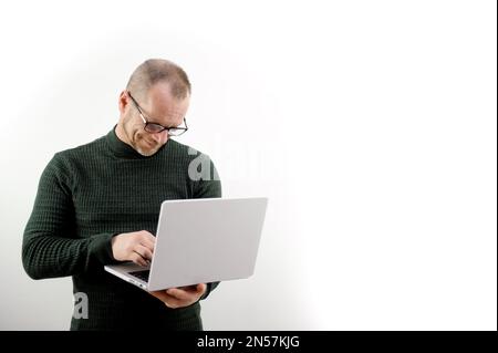 Focused man in glasses using a laptop, typing on the keyboard, writing an email or message, chatting, shopping, a successful freelancer working online on a computer stands on a white background Stock Photo
