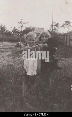Vintage Photograph of a  young Girl and her pet Dog / hand holding puppy / hugging dogs / Dog Hugs / little sisters posing for the camera at the 1930s. one of them is holding her little black dog / black puppy. / Holding dog under arm Stock Photo