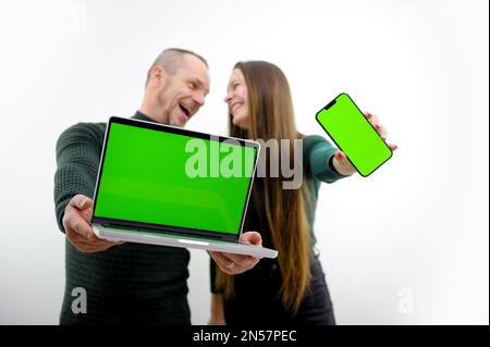 Use this new app. Cheerful couple point to smartphone and laptop with blank screen, recommend mobile app, advertise your website, mockup for design. chroma key green screen selective focus Stock Photo
