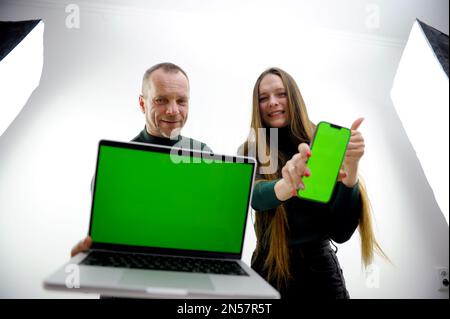 Use this new app. Cheerful couple point to smartphone and laptop with blank screen, recommend mobile app, advertise your website, mockup for design. chroma key green screen selective focus Stock Photo