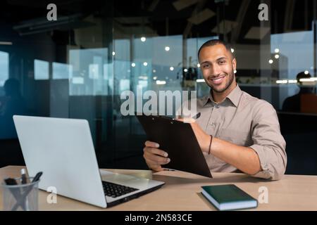 Portrait of smiling and satisfied businessman with achievement results, african american paper work smiling and looking at camera, young financier writing document inside office using laptop and pen. Stock Photo