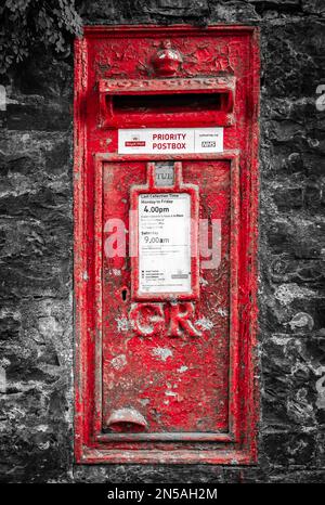 An old bright red wall-set postbox against a black and white background. The postbox dates from King George V, so peeling paint shows its age Stock Photo