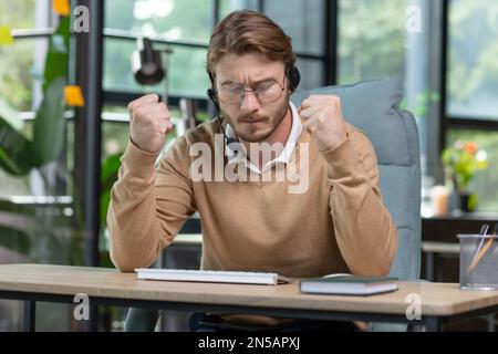 Angry office worker working inside office, young businessman in casual clothes with headset for video call, male nervous tech support worker and customer consultation online. Stock Photo
