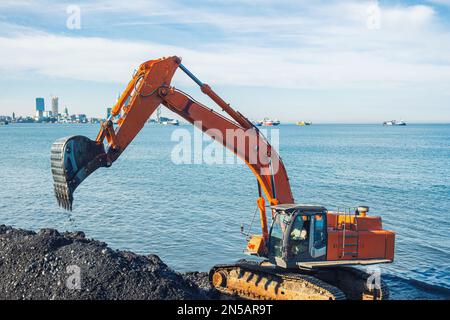 Excavator or digger working on earthmoving at shore protection works in Batumi. Orange backhoe digs sand and gravel in quarry. Digger during excavatio Stock Photo
