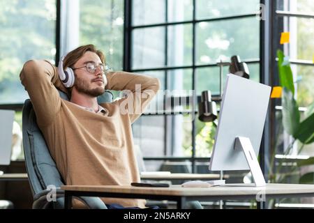 Young successful man in office resting with eyes closed and hands behind head, programmer finished work satisfied with achievement results and listening to music in headphones and podcast online. Stock Photo