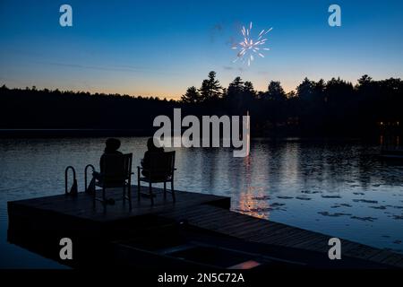 A silhouette of two women sit on a pier at a lake and watch fireworks explode in the night sky on the Fourth of July holiday. Stock Photo
