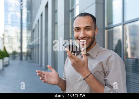 Outdoor portrait of young smiling African American man outside office building, man recording audio message smiling and looking at camera, businessman satisfied with work of AI voice assistant. Stock Photo