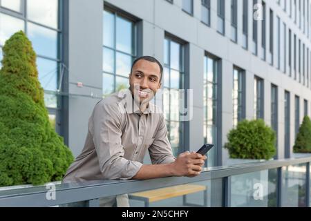 Cheerful young student in shirt using phone outside university campus, african american man smiling and looking away, holding smartphone, typing message and making call. Stock Photo