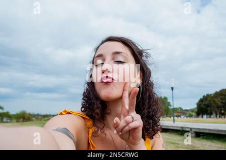 Selfie portrait of young Argentinian Latina woman, white-skinned with red lips and dressed in yellow, outdoors looking at the camera giving a kiss and Stock Photo