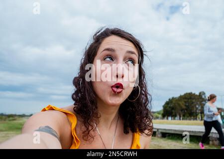 selfie portrait of young latin argentinian woman, doubting surprised with open mouth and looking up, red lips, yellow dress, cloudy sky in background Stock Photo
